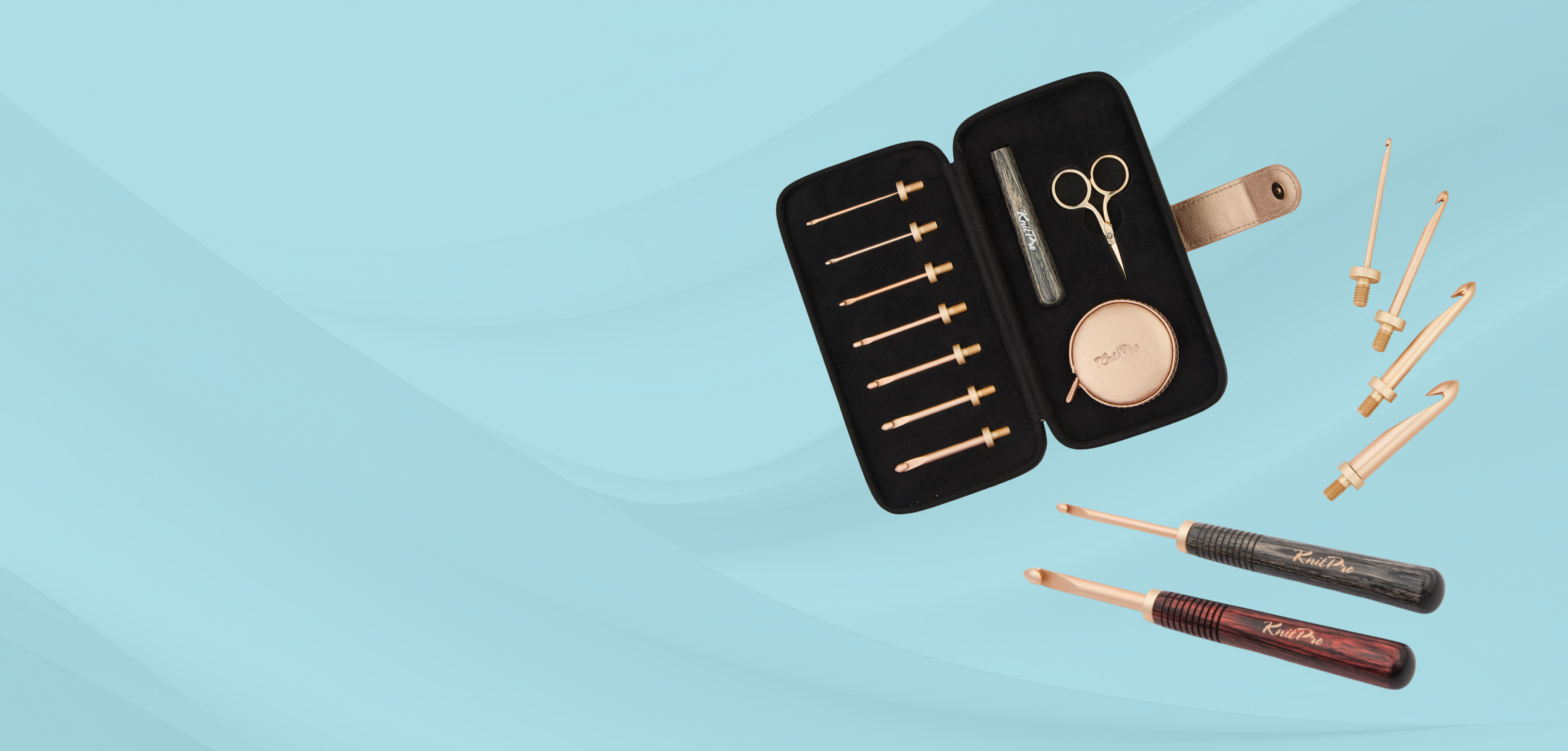 Set of rose gold crochet hooks and tools including scissors and a tape measure in a black case on a light blue background